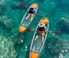 Two people in transparent kayaks paddle over clear sparkling water revealing the rocky seabed below