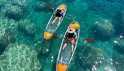 Two people in transparent kayaks paddle over clear, sparkling water revealing the rocky seabed below.