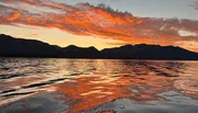 A fiery sunset paints the sky and reflects on the rippled surface of a lake against a backdrop of dark mountains.