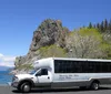 A tour bus with Discover Lake Tahoe Scenic Tours printed on its side is parked near a lakeshore with a rocky outcrop in the background under a clear blue sky