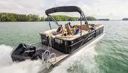 A group of people are enjoying a ride on a modern pontoon boat equipped with a Mercury outboard engine, gliding across a tranquil lake.