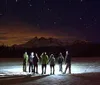 A group of people stand on a snow-covered ground at night gazing at a starry sky with a backdrop of mountains