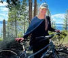 A person is standing with a bicycle in a forested area smiling at the camera with a lake visible in the background