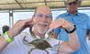 A smiling man is playfully holding a crab in front of him as if it's pinching his chin while another man observes in the background.