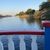 The image depicts a tranquil river scene viewed through a railing with white balusters and a red and blue border, under a clear sky.