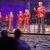A group of performers in matching red jackets adorned with gold patterns are singing on stage in front of an audience.