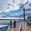Passengers enjoy the view from the deck of a riverboat under a sky scattered with clouds.