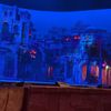 The image shows a theater stage set up to resemble an exotic, stylized town bathed in blue light, likely before or during a performance.