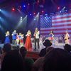 Performers are on stage singing in front of a large American flag backdrop with an audience in the foreground.