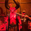 A person poses beside a large demonic figure with towering horns as part of a macabre and dramatic display, complete with skulls on a table in the foreground.