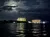 A nighttime cityscape is illuminated by the glow of buildings on the shore, with reflections shimmering on the water under a cloud-veiled moon.