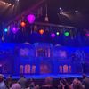 The image shows a vibrant theater stage with a colorful, ornate set design and hanging lanterns, with an audience in the foreground possibly waiting for a performance to begin.