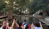 Tourists on a boat tour are capturing photos of a scenic riverbank lined with trees and walkways.