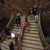 Visitors are ascending and descending a grand wooden staircase with ornate decorations in a lavish interior setting.
