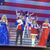 Performers in patriotic attire are singing and entertaining on stage, set against a large American flag backdrop.