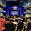 An audience is seated at tables in a theater with their attention directed towards a brightly lit stage framed by a decorative arch with the words 