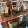 The image shows a museum display with three large, weathered silver ingots recovered from the SS Gairsoppa shipwreck, and a viewer observing them from behind the glass casing.