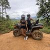 Two people wearing helmets are sitting on an all-terrain vehicle on a dirt track with a forested, mountainous backdrop.