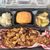 The image shows a barbecue meal featuring pulled pork, baked beans, macaroni and cheese, a dinner roll, and apple cobbler, all served in a black plastic takeout container.