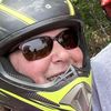 Two people wearing motorcycle helmets are smiling for a selfie outdoors, with one wearing a red helmet and the other a yellow and black one.