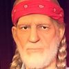 The image shows a life-sized figure, resembling an older man with braided hair and wearing a red cap, a plaid shirt, and jeans, staged in front of a red curtain alongside an informational display and a musical instrument, likely a waxwork or a statue, designed to mimic a person's appearance.