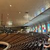 The photo shows the interior of a spacious auditorium or theater with rows of wooden seats, colorful stained glass windows, and a high wooden ceiling with mounted lights.