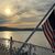 The image captures a serene sunset over a body of water with a gently waving American flag in the foreground, framed by an ornate white railing.