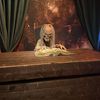 A spooky animatronic figure with a skeleton-like face reads a book at a wooden desk against a backdrop with a cosmic or mystical theme.