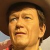 A wax figure of a cowboy, complete with a hat, bandana, and rifle, stands against a backdrop of a desert scene with a cactus.