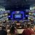 Audience members are seated at tables in a multi-tiered theater, looking towards a brightly lit stage with 