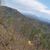 The image captures a scenic mountainous landscape viewed from behind the seat of an off-road vehicle, with a clear blue sky above and trees beginning to show spring growth.