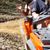 Two individuals are riding an orange ATV on a dirt trail with leafy trees in the background, and the passenger gestures while wearing a motorcycle helmet.