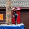 A person in a plaid shirt and jeans is engaged in a log climbing activity, using a rope and climbing spikes to ascend a tall pole, with a padded safety base below.