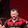 Two individuals dressed in lumberjack attire are preparing for a wood chopping activity on a stage, overlooked by another person, against a backdrop with playful, themed signage.