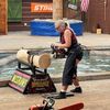 A person dressed in a plaid shirt and protective gear is operating a chainsaw on a log in a lumberjack show setting.