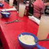 The image depicts a casual dining setting with a bowl of what appears to be clam chowder and a lemonade-like beverage on a red table, with blurred background activity indicating other diners.