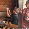 A young boy is aiming a toy gun with concentration while an adult woman watches him, both standing next to a wooden rack with various other replica guns.