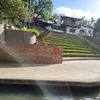 The image shows a sunny outdoor setting with a canal or river in the foreground, a paved embankment, and a grassy, stepped area leading up to white buildings with a sign saying 