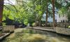 The image shows a serene urban river walk flanked by lush green trees, with sunlight filtering through the foliage.