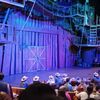 The image shows an audience seated at tables inside a theater with a rustic barn stage set, waiting for a performance to begin, as some attendees wear matching white cowboy hats.