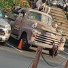 The image shows a vintage truck with a large sculpture of a cow on its bed, parked among other vehicles in a sunny outdoor setting.