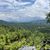 The image captures a verdant mountainous landscape under a partly cloudy sky, with hints of human habitation tucked among the foliage and the corner of an off-road vehicle visible in the foreground.