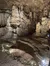 The image shows a dimly lit cavern with intricate stalactite and stalagmite formations, illuminated by artificial lights enhancing the textures and details of the cave's interior.