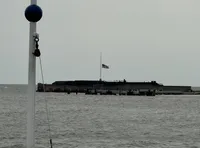 An overcast sky looms over a body of water with a half-mast American flag flying at a distant building, viewed past a pole with a spherical buoy attached at the top.