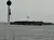 An overcast sky looms over a body of water with a half-mast American flag flying at a distant building, viewed past a pole with a spherical buoy attached at the top.