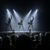 The image shows a live drumming performance on a stage with dramatic backlit lighting, captivating an audience sitting in the darkened theater.