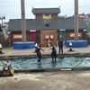 Three people are standing in a lumberjack-themed water arena with one person apparently in mid-performance, showcasing an event likely part of a lumberjack show.