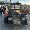 An old, rustic car decorated with various items and signs is parked in a lot while people walk by.