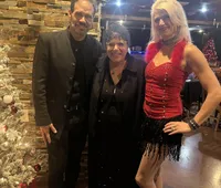 Two men and a woman are smiling for a photo next to a Christmas tree, with the woman wearing a festive red outfit and the men in formal black attire.