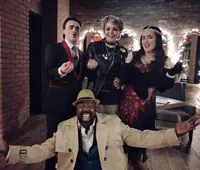 A group of four people, dressed in vintage and stylish attire, poses enthusiastically in a warmly lit room with exposed brick walls and twinkling lights.